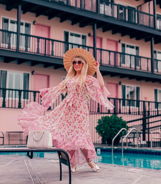 blogger lizzie in lace wearing a floral dress with ruffled cascading sleeves and pink on a white background. She's standing by a pool in front of a pink hotel. 