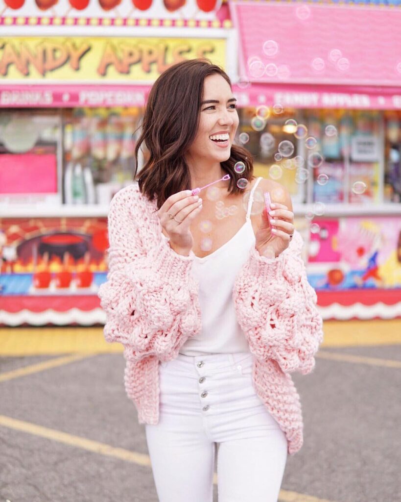 blogger wearing the pink bell sleeve chunky knit sweater with pom pom balls on the sleeves. the blogger is wearing it over a white tank top and jeans and standing in a carnival blowing bubbles