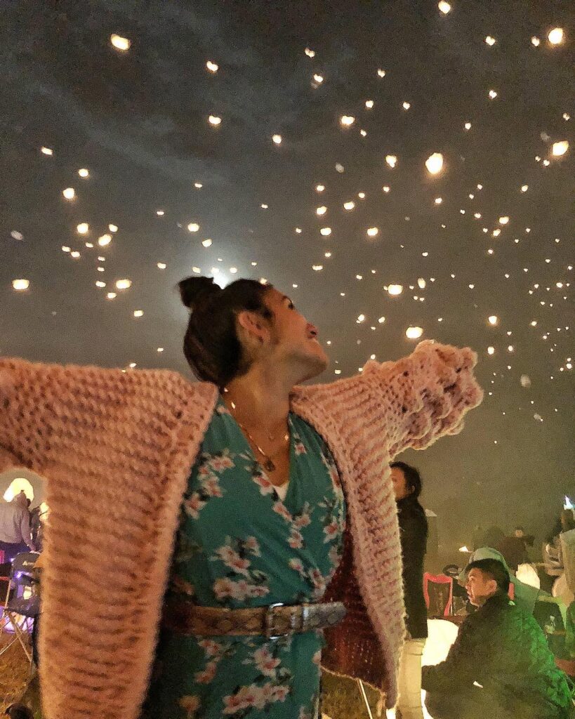 blogger wearing the pink bell sleeve chunky knit sweater with pom pom balls on the sleeves. the blogger is standing at a festival at night with lights in the sky