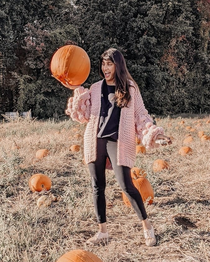 blogger wearing the pink bell sleeve chunky knit sweater with pom pom balls on the sleeves standing in a pumpkin patch