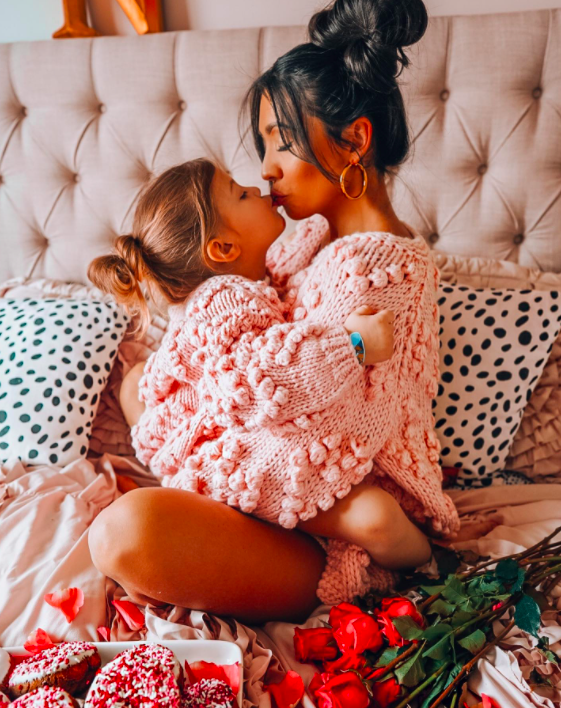 a mom and her daughter sitting on a bed wearing the matching pink mommy-and-me sweaters