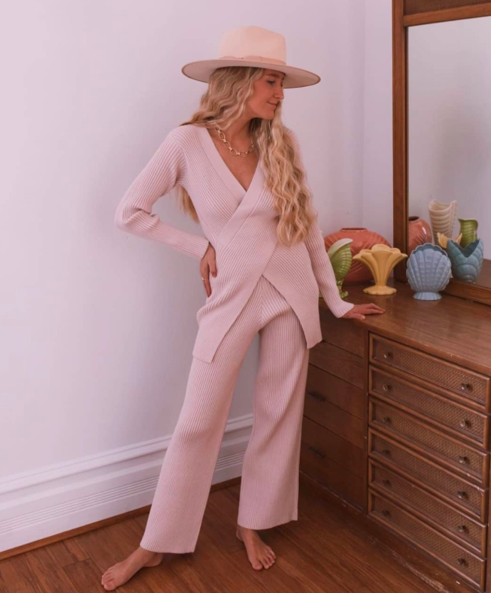 chicwish blogger wearing the long-sleeve knit sweater and knit pants set in beige with bare feet standing beside a brown wood dresser and wearing a hat